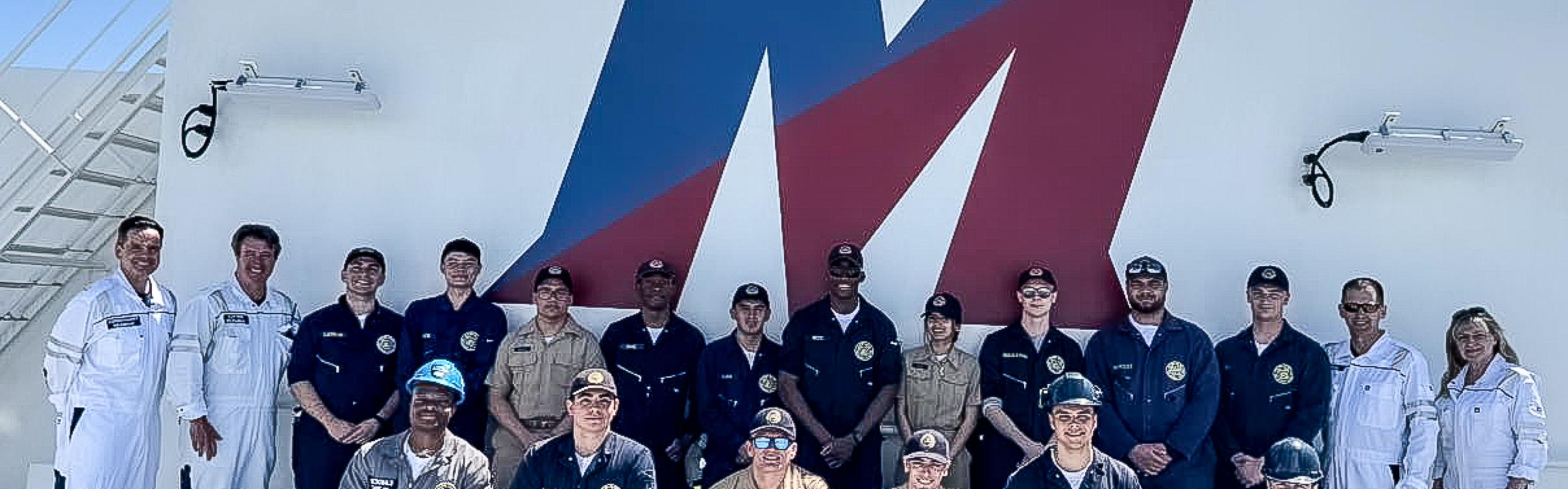 Group photo of Maritime College cadets, crew, and staff in uniform standing and kneeling on the deck of the Empire State VII training ship, posed in front of a large red, white, and blue "M" emblem under a clear blue sky.